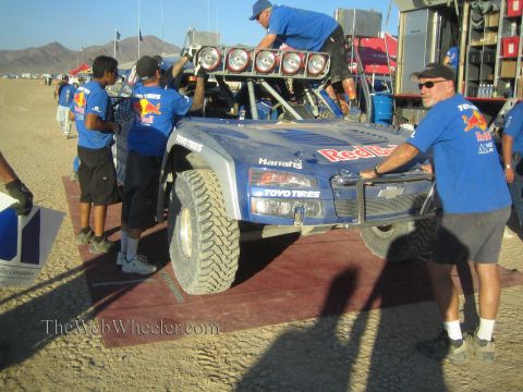 1990 Ford Bronco - crew in blue Red Bull shirts services a blue Chevrolet trophy truck with roof-mounted lights in a dusty desert pit lined with red mats.