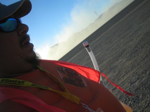1990 Ford Bronco - man in a cap and mirrored sunglasses leans into a close selfie, wearing a red BFGoodrich shirt with a hydration tube against a dusty desert…