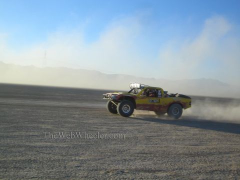 1990 Ford Bronco - yellow off-road race truck races across a flat desert lakebed at speed, trailing a large cloud of dust against distant mountains.