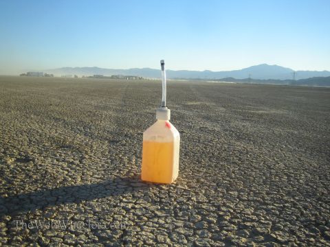 1990 Ford Bronco - white plastic jug holding orange liquid and topped with a tall antenna marker stands on a cracked dry lakebed with mountains on the horizon.