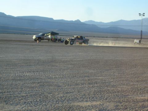 1990 Ford Bronco - open off-road buggy kicks up dust as it races past a parked white pickup and pit canopy on a flat desert lakebed below mountains.