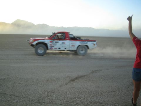 1990 Ford Bronco - white and red off-road race truck blasts across a dusty desert track while a woman in a red shirt raises a thumbs-up in the foreground.