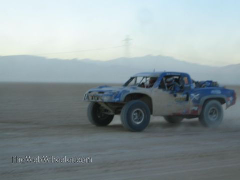 1990 Ford Bronco - blue and white off-road race truck powers across a flat desert lakebed at speed, raising dust under a hazy sky with power lines behind.
