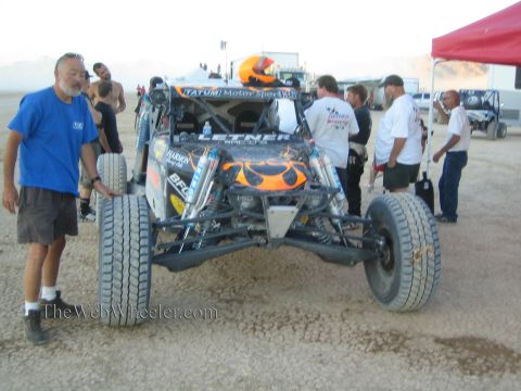 1990 Ford Bronco - orange flame-painted tube-chassis off-road race car sits in a desert pit surrounded by crew, a helmeted driver, and pop-up canopies.