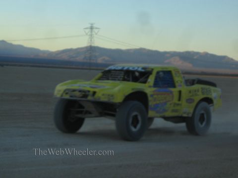 1990 Ford Bronco - yellow off-road race truck races across a desert lakebed at speed, with a transmission tower and mountains in the background at dusk.