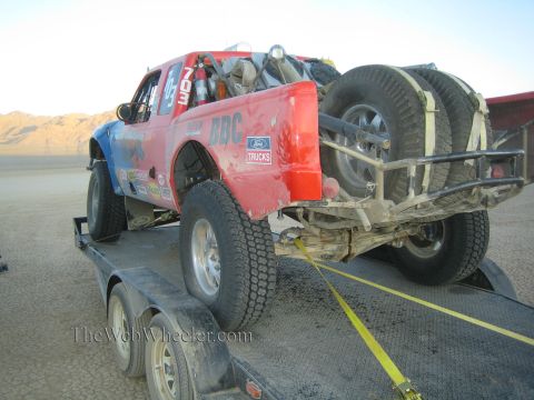 1990 Ford Bronco - red and blue Ford off-road race truck, carrying mounted spare tires, sits strapped onto a tandem-axle flatbed trailer on a desert lakebed a…
