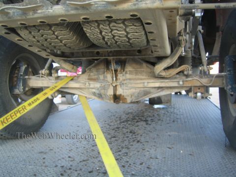 1990 Ford Bronco - low-angle underside view shows the solid rear axle, differential, and leaf springs of a dirt-caked off-road truck strapped down on ramps.