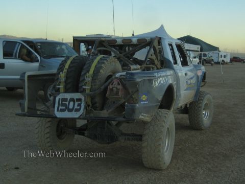 1990 Ford Bronco - gray tube-chassis off-road race truck numbered 1503 with twin rear-mounted spare tires sits in a desert pit among canopies and haulers at d…