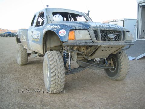 1990 Ford Bronco - mud-caked blue Ford prerunner race truck with a tubular front end and long-travel suspension sits in a dusty desert pit area.
