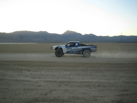 1990 Ford Bronco - blue and white desert race truck races across a dry lakebed at dusk, kicking up a trail of dust against distant mountains.