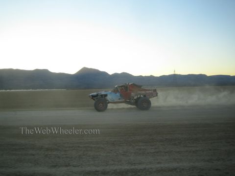 1990 Ford Bronco - off-road race truck speeds across a dusty desert flat at dusk with mountains silhouetted behind it.