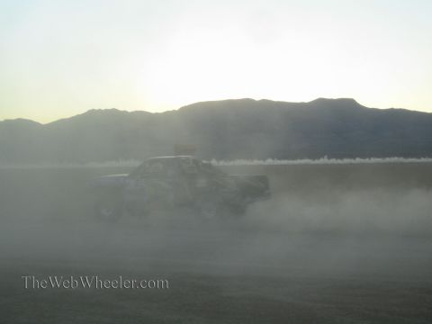 1990 Ford Bronco - race vehicle barrels through a thick dust cloud on a desert flat at dusk, nearly hidden by the haze.