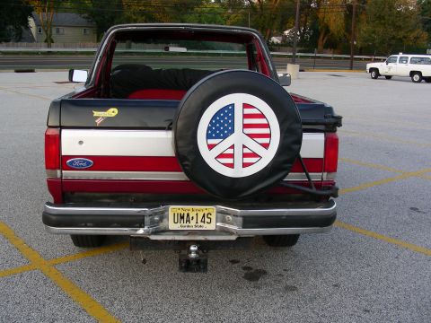 1990 Ford Bronco - This Ford Bronco features a patriotic peace sign on its spare tire cover, parked in a lot with another vehicle visible.