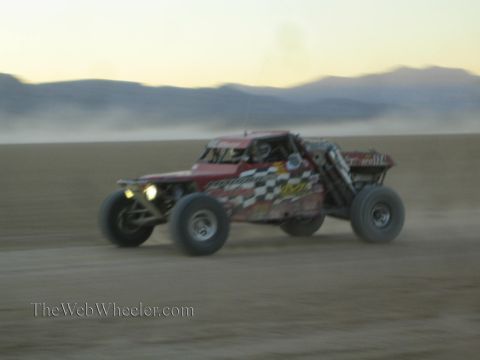 1990 Ford Bronco - red tube-chassis off-road race buggy with headlights blazing races across a desert flat at dusk.