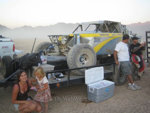 1990 Ford Bronco - woman and young girl crouch near a silver and yellow number 118 off-road race truck loaded on a trailer in a dusty desert pit.