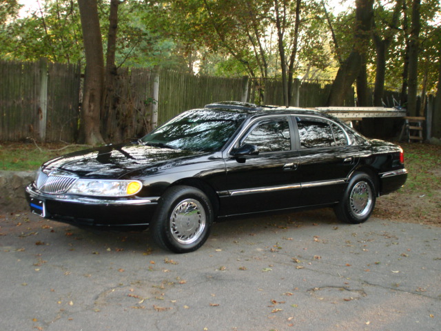Black Lincoln Continental parked on a paved surface with trees and a wooden fence in the background