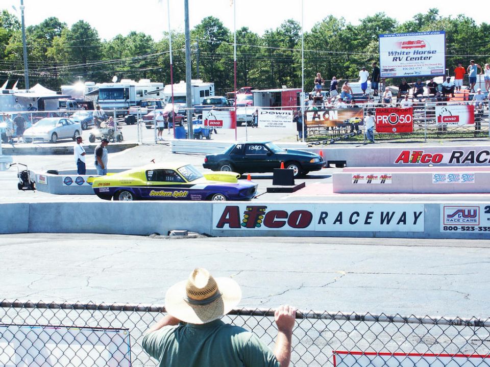 Drag strip with a yellow and purple drag car and a dark sedan, spectators in bleachers, and RVs in the background