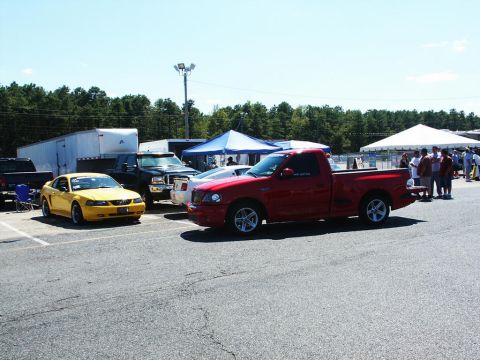 2001 Mercury Grand Marquis - A red pickup truck and a yellow coupe are parked at a car show with tents and trees in the background.
