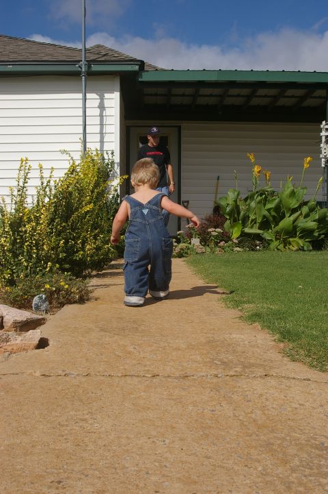 1998 Jeep TJ - A young child in denim overalls walks down a sidewalk towards a house, with another person visible in the doorway.
