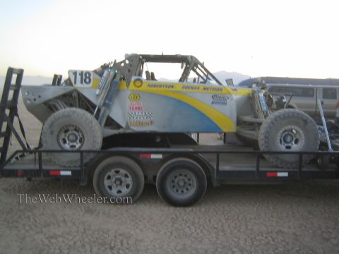 1990 Ford Bronco - Side profile of a silver and yellow number 118 off-road race buggy strapped to a dual-axle flatbed trailer in the desert.