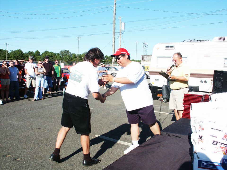Two men shaking hands at a car show, with a crowd and vehicles in the background