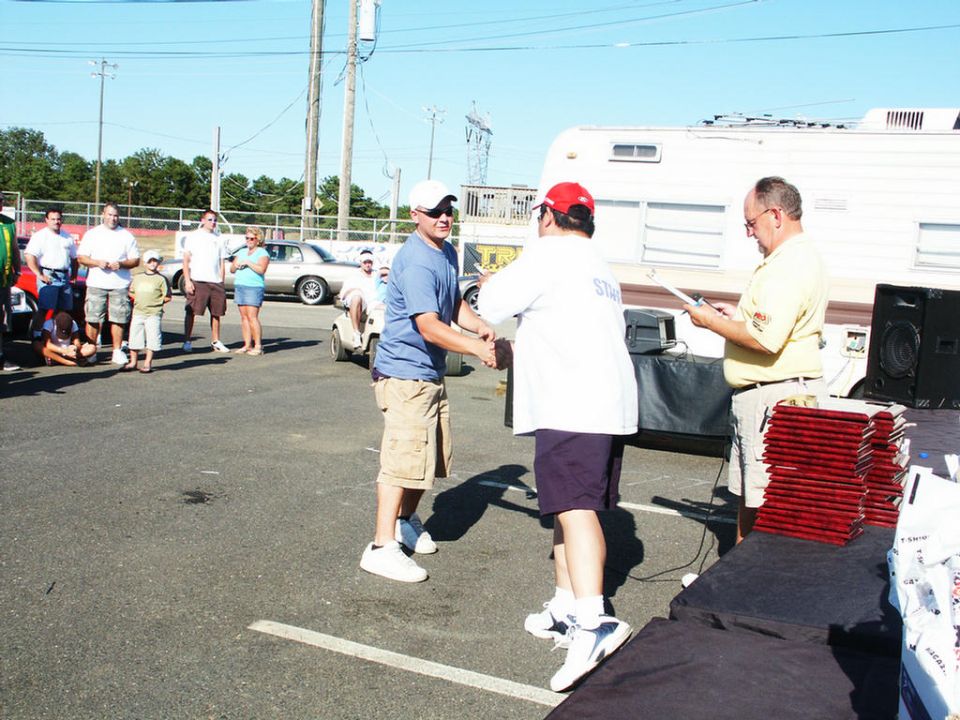 Two men shaking hands at an outdoor event, with a white trailer and several cars visible