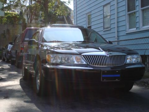 1997 Ford Crown Victoria - A black Lincoln Continental is parked in front of a blue house, with other vehicles lined up behind it.