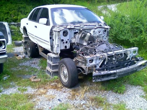 2002 Ford Crown Victoria - A white sedan, stripped of its front end and engine components, is propped up on cinder blocks in a grassy area.