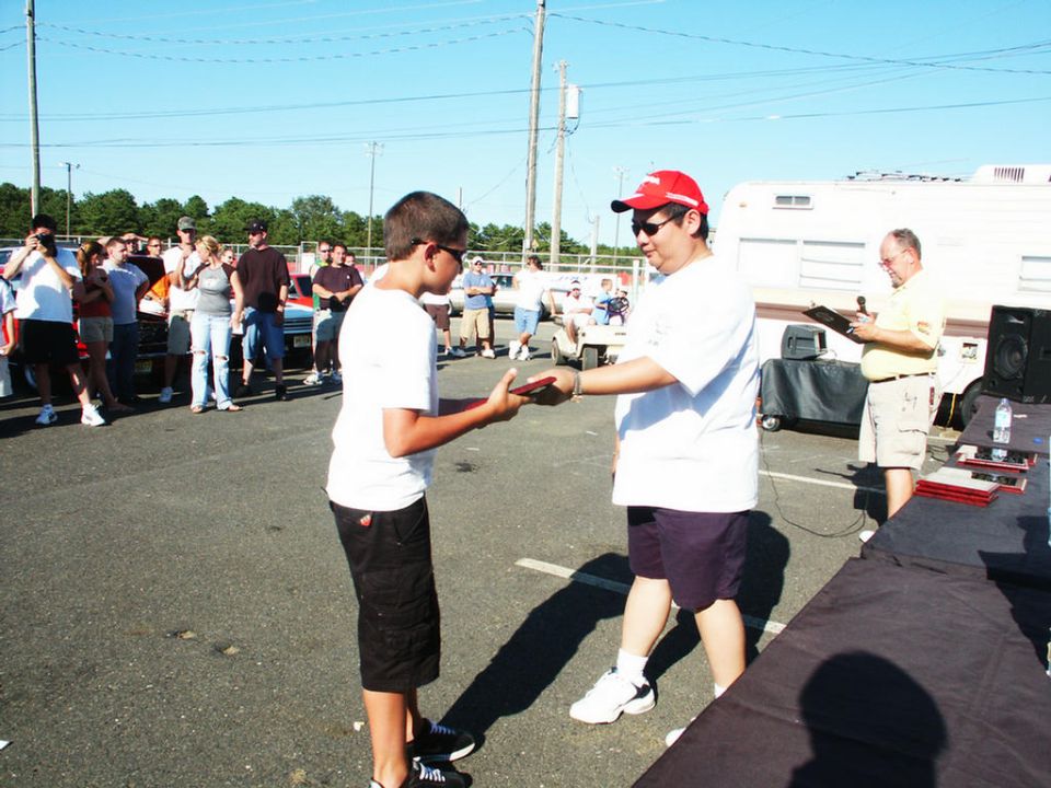 Two boys receiving awards at a car show with other people and vehicles in the background