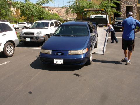 1992 Ford Taurus - A blue 1992 Ford Taurus is loaded onto a tow truck in a parking lot.