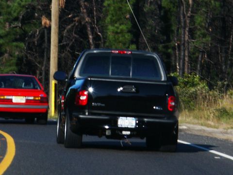2001 Mercury Grand Marquis - A black pickup truck is seen from behind, driving on a road next to a red car.