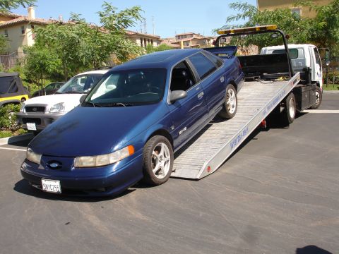 1992 Ford Taurus - A blue sedan is being loaded onto a tow truck, with other vehicles visible in the background.