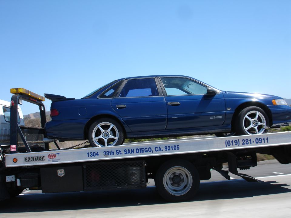 Blue sedan with a spoiler on a tow truck, side view. Text on tow truck reads '1304 38th St. SAN DIEGO, CA. 92105'