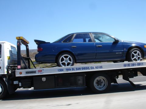 1992 Ford Taurus - A blue 1992 Ford Taurus sedan, equipped with a rear spoiler, is being transported on a tow truck.