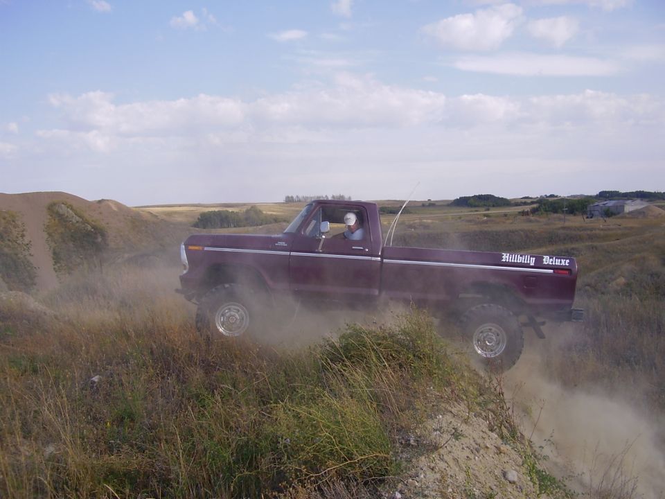 1979 Ford F-250 pickup truck with 'Hillbilly Deluxe' decal on the side, kicking up dust while driving on a dirt hill
