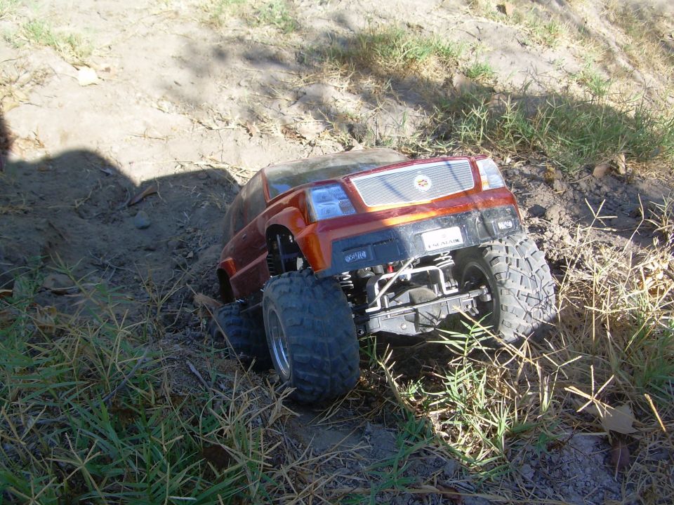 Red Escalade-body RC crawler nosed into a dirt rut surrounded by dry grass and a shadow