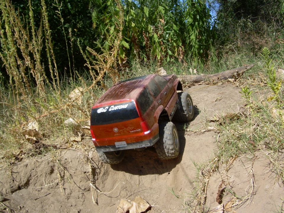 Rear three-quarter of red B&C Customs RC Escalade crawler on sandy slope by green foliage