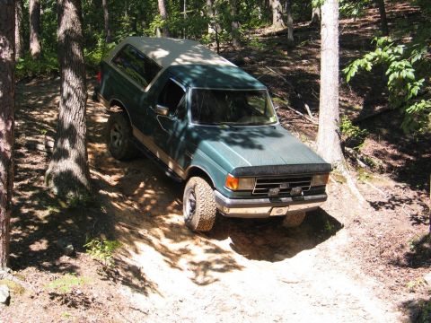 1990 Ford Bronco - A green Ford Bronco navigates a wooded dirt trail, showing its front 3/4 view with a tan top.