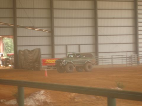 1989 Ford Bronco - A green SUV with large tires is seen driving on a dirt surface inside a large building.