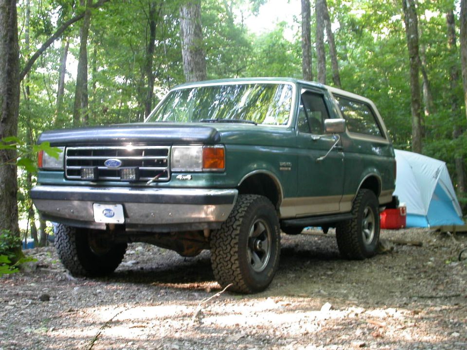 1990 Ford Bronco parked in a wooded area with a tent and cooler nearby