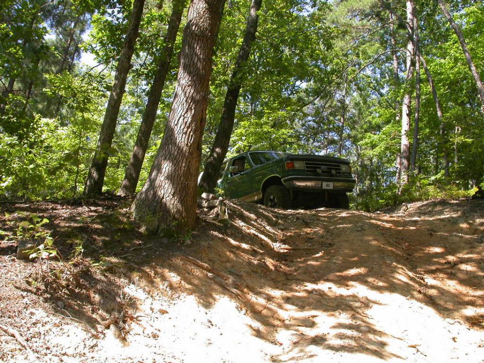 Green Ford Bronco climbing a dirt hill in a wooded area. The front of the vehicle is visible