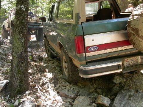 1990 Ford Bronco - Two off-road vehicles navigate a challenging rocky path through a wooded area.