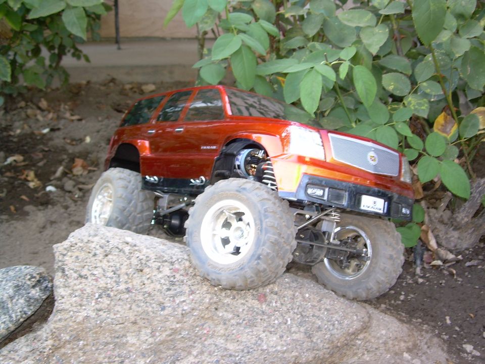Orange-red Escalade RC crawler passenger side cresting a granite boulder near green leaves