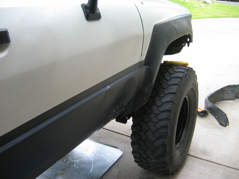 White truck raised in driveway with wheel removed, mud-terrain tire leaning on body and inner fender liner off