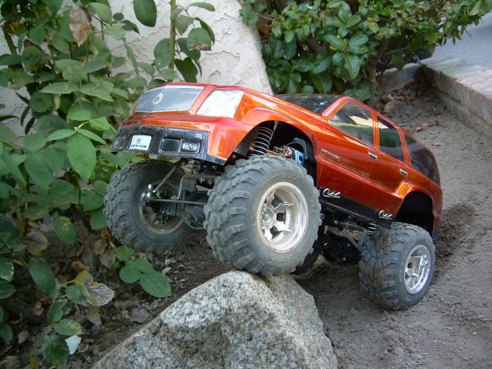 Orange Escalade RC crawler descending a boulder, exposed shocks and chassis, brush behind