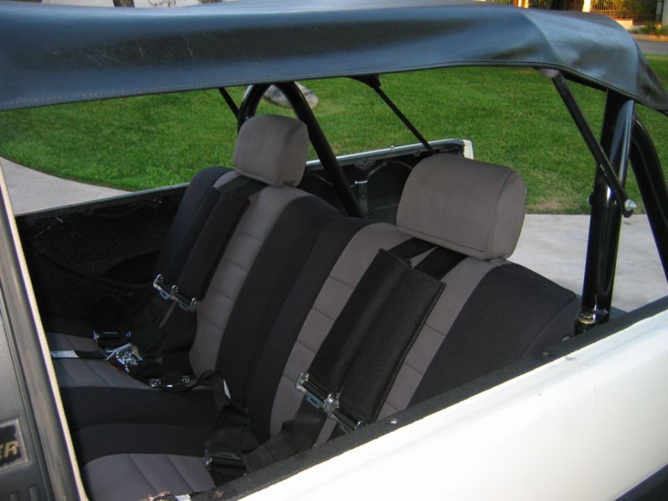 Interior of a vehicle showing two rows of seats with black and grey upholstery and racing-style seatbelts. A black roll cage is visible