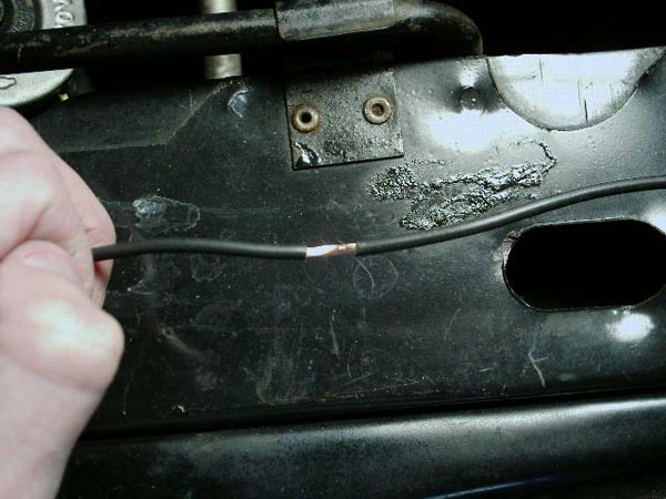 Hand holding a black wire with exposed copper strands against black metal bodywork with an oval hole
