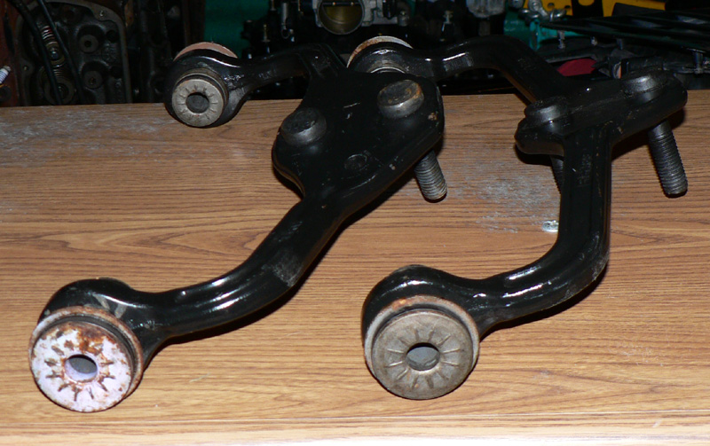 Two black control arms with bushings, one bushing rusted, arranged on wood bench