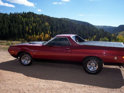1972 Ford Ranchero - A red 1972 Ford Ranchero is parked on a dirt road, with a backdrop of a wooded hillside under a blue sky.