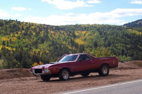 1972 Ford Ranchero - A maroon 1972 Ford Ranchero is parked on a dirt road, with a backdrop of a colorful, tree-covered hillside under a partly cloudy sky.